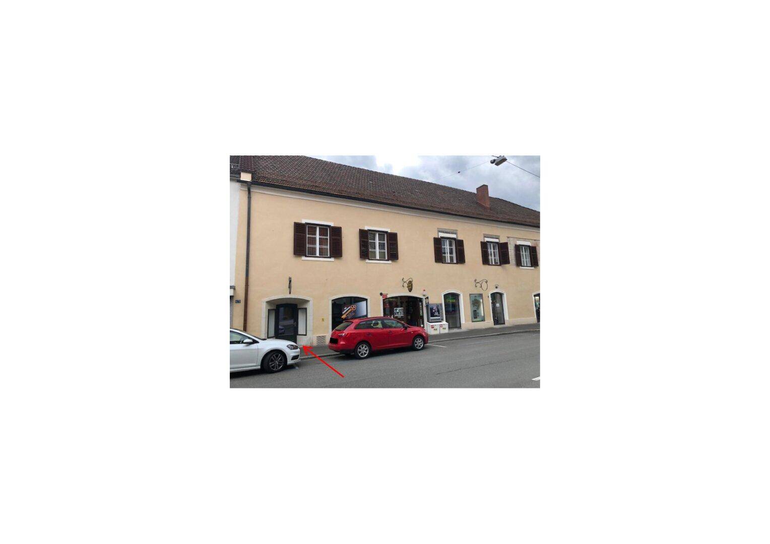 Fassade eines Gebäudes von der Straße aus gesehen, mit geparkten Autos und einem Eingangspfeil.