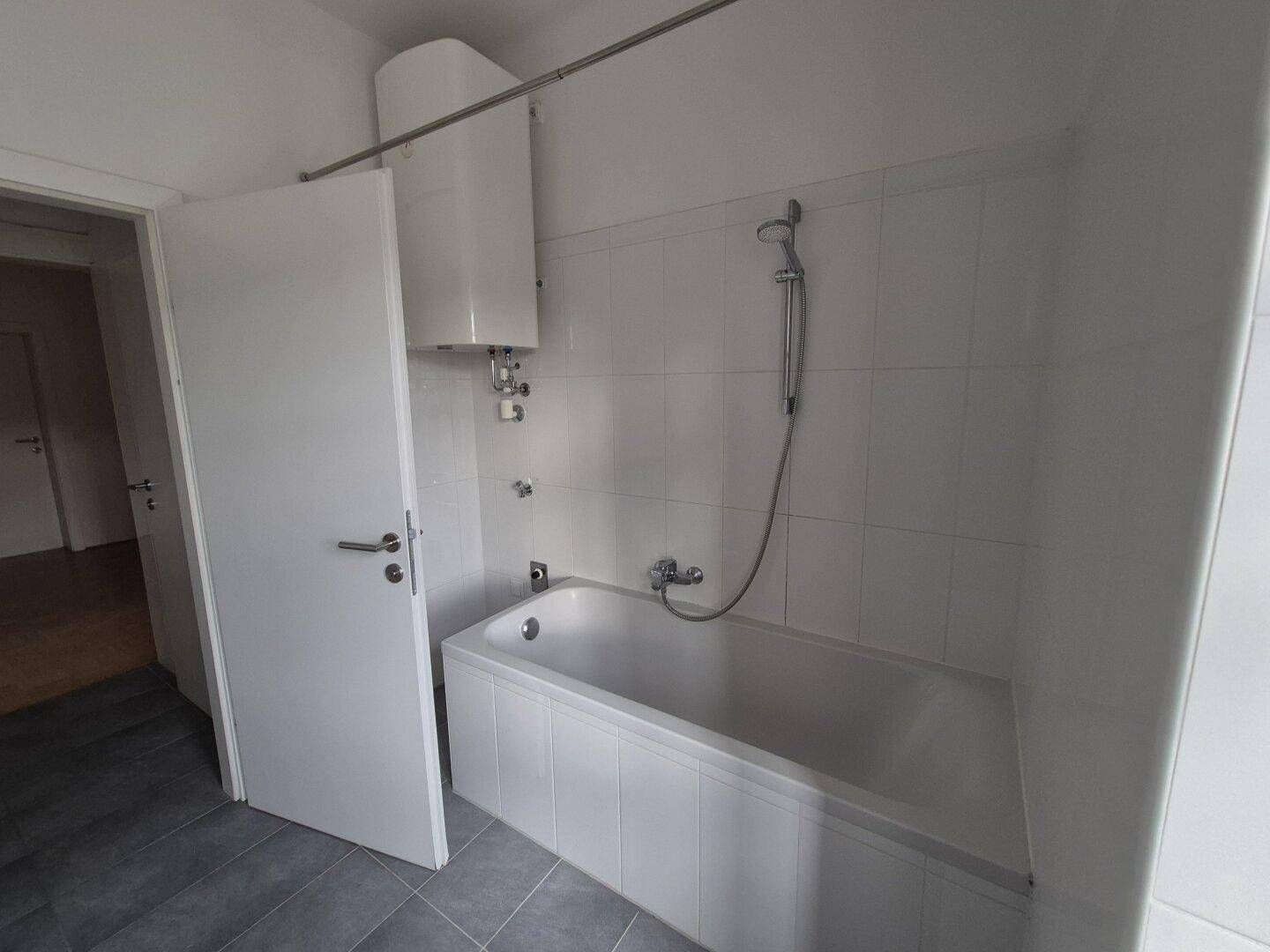 Badezimmer mit Badewanne, weißen Fliesen und einem Warmwasserbereiter an der Wand.