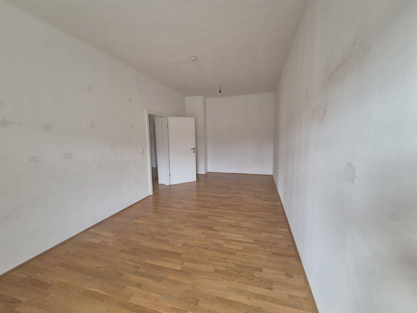 Geräumiges, leeres Zimmer mit Holzboden und einer offenen Tür zu einem weiteren Bereich.