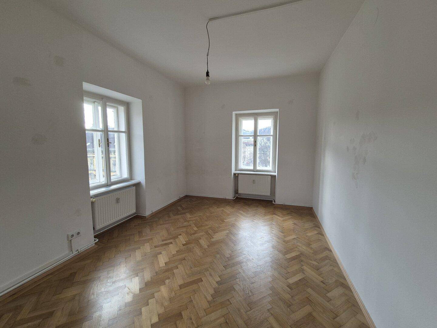 Helles Zimmer mit Fischgrätparkettboden und zwei Fenstern, die viel Tageslicht hereinlassen.