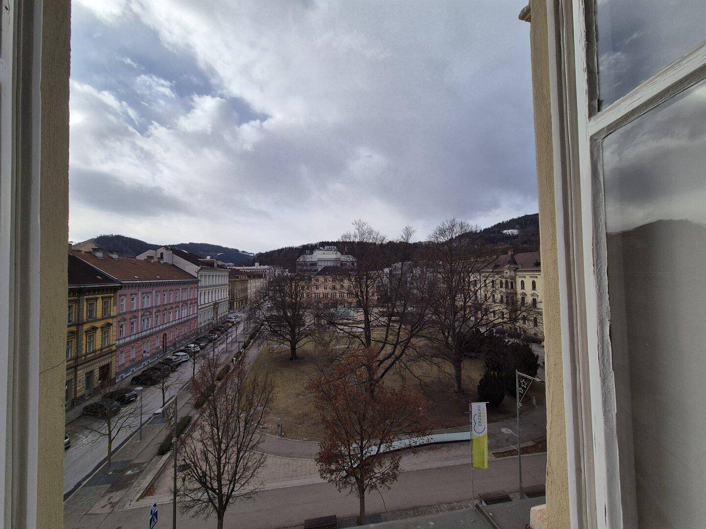 Blick aus dem Fenster auf einen Stadtplatz mit Bäumen und umliegenden Gebäuden.