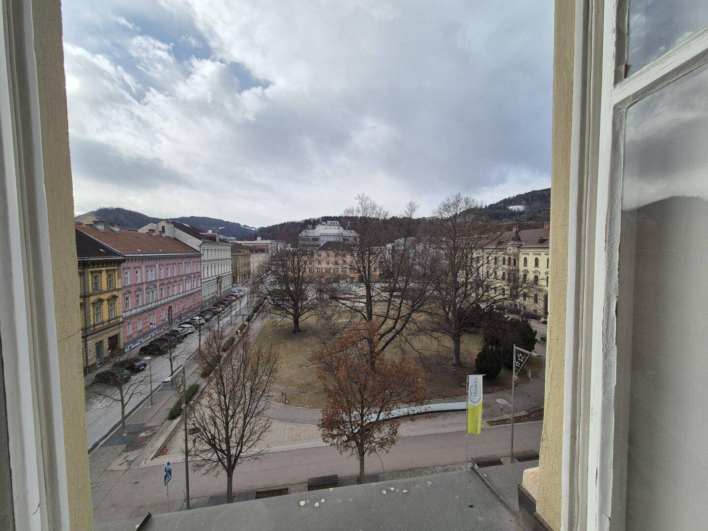 Weitblick aus dem Fenster auf einen belebten Stadtplatz mit historischen Gebäuden und Hügeln im Hintergrund.