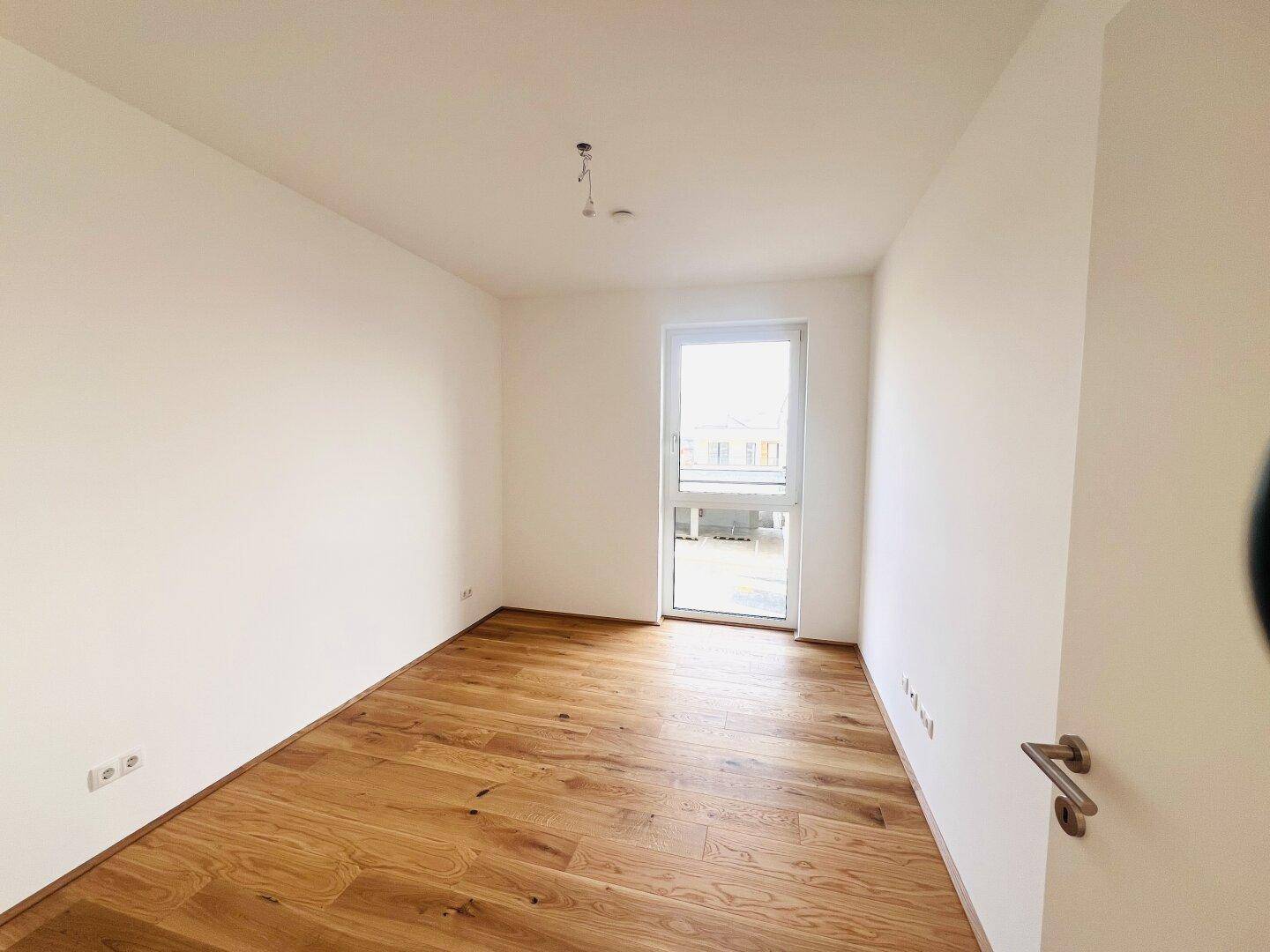 Leeres Zimmer mit Parkettboden und einer Tür, die zu einem kleinen Außenbereich führt.