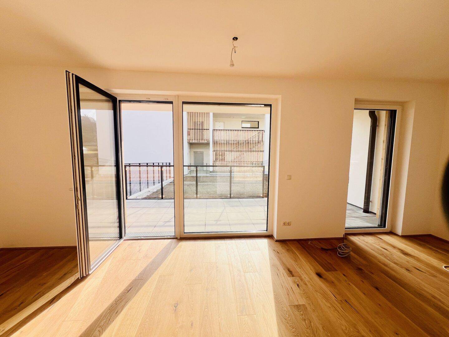 Helles, leeres Zimmer mit Holzboden und großen Glasfronten, die zu einem Balkon oder einer Terrasse führen.