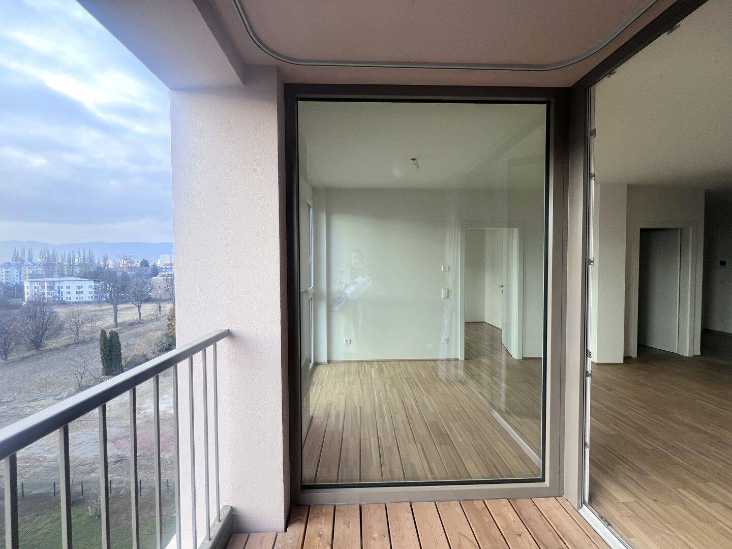 Blick vom Balkon mit Holzbelag auf die Umgebung und den hellen, modernen Innenraum.