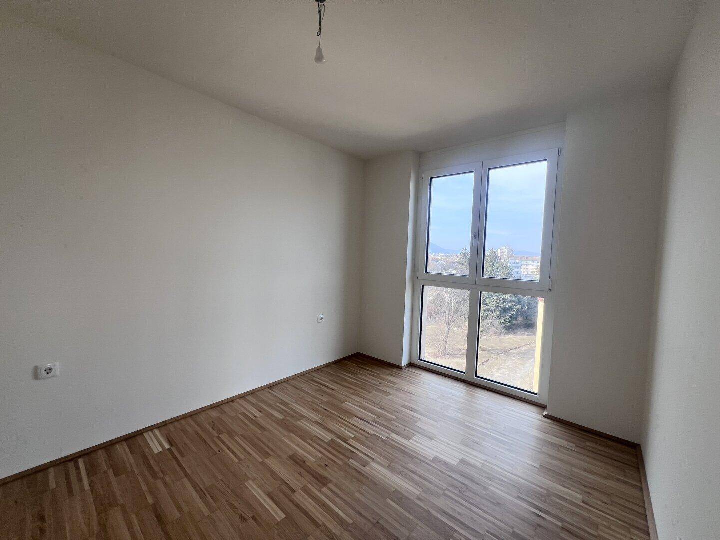 Helles Schlafzimmer mit Parkettboden und großem Fenster mit Blick ins Grüne.