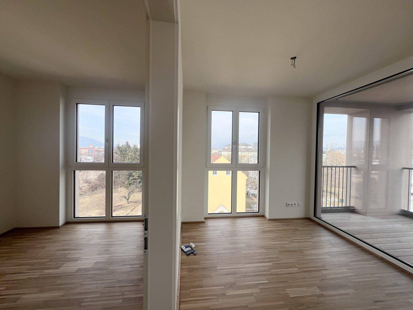 Geräumiger Wohnbereich mit Parkettboden, großen Fenstern und Zugang zum Balkon.