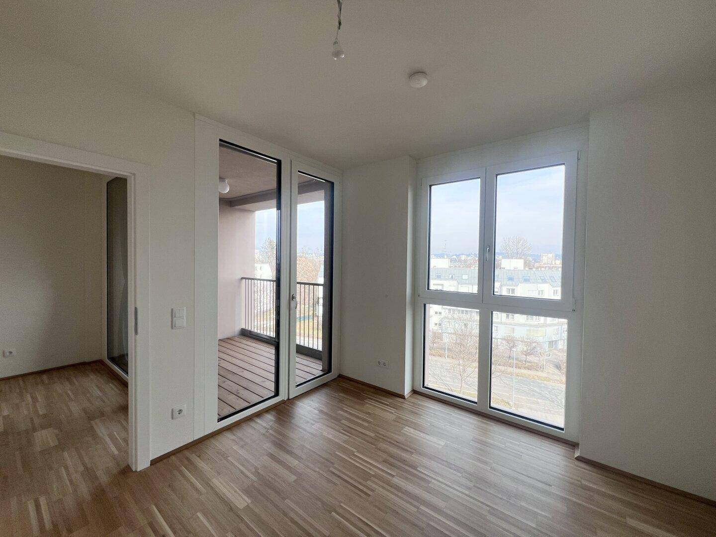 Lichtdurchfluteter Raum mit Parkettboden, großen Fenstern und direktem Zugang zum Balkon.