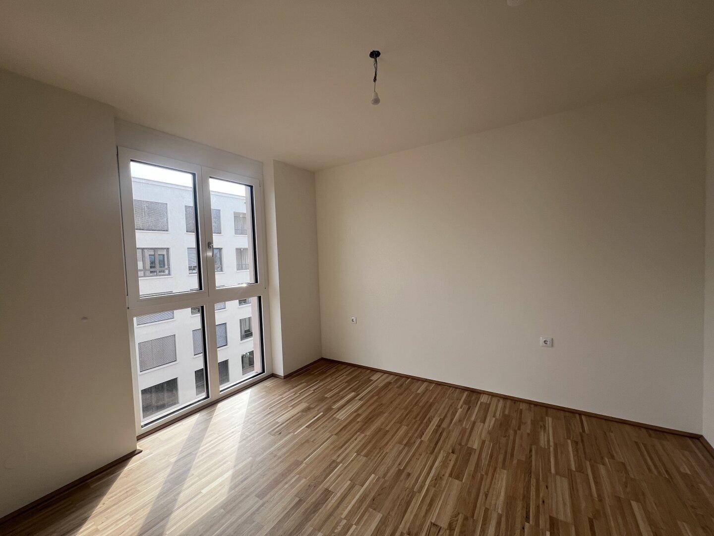 Leeres Zimmer mit Parkettboden und großen Fenstern, die viel Tageslicht hereinlassen und eine helle Atmosphäre schaffen.