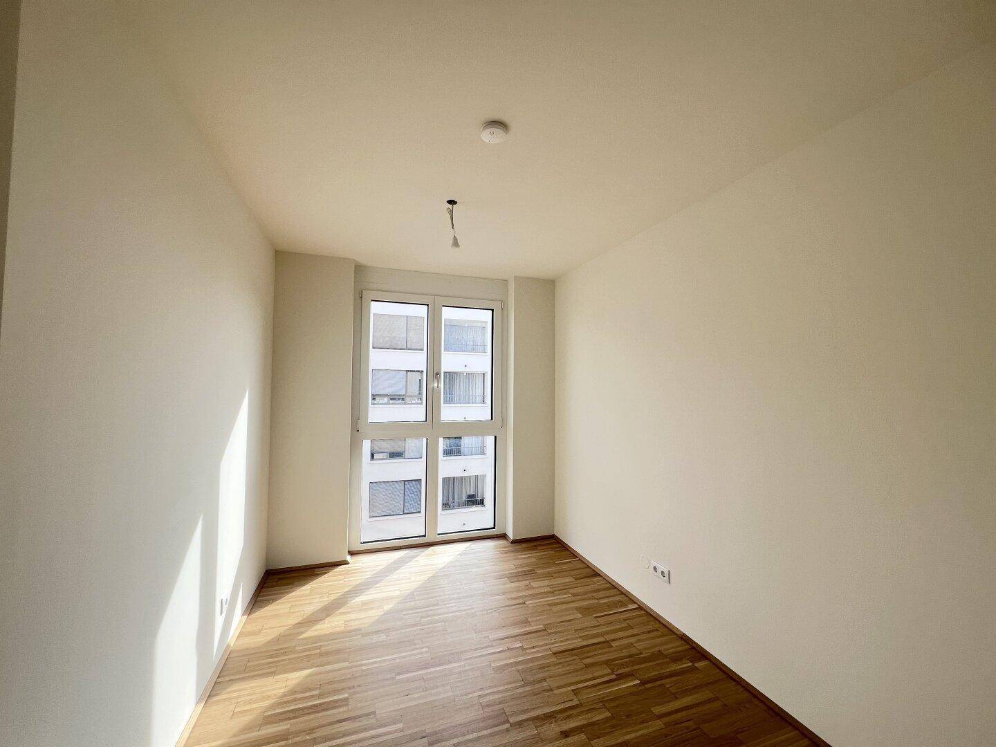 Heller, leerer Raum mit Parkettboden und großem Fenster, das viel Tageslicht hereinlässt.