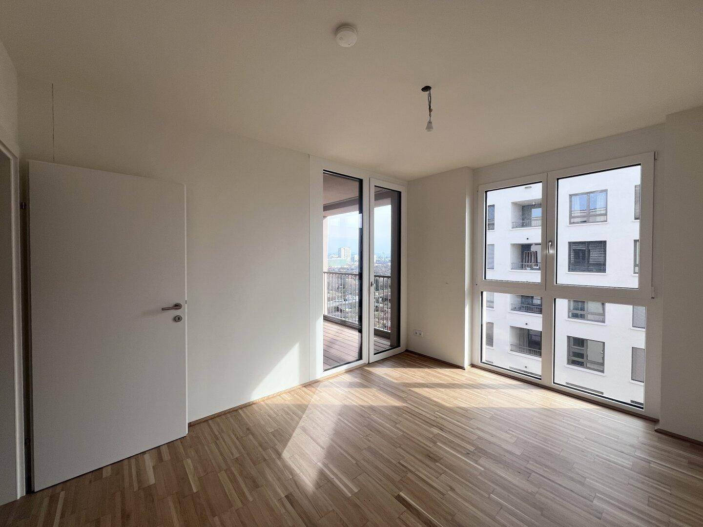 Lichtdurchfluteter Raum mit Holzboden und Zugang zu einem Balkon sowie Blick auf die Stadt.