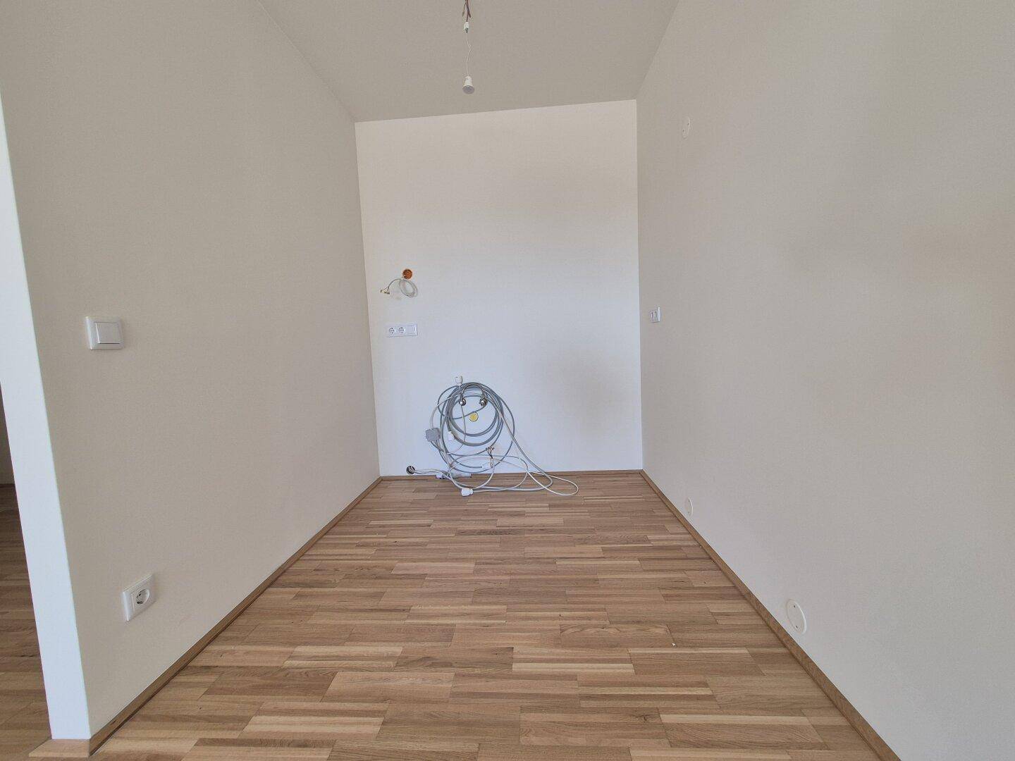 Leerer Raum mit Parkettboden und vorbereiteten Anschlüssen für eine Küche.