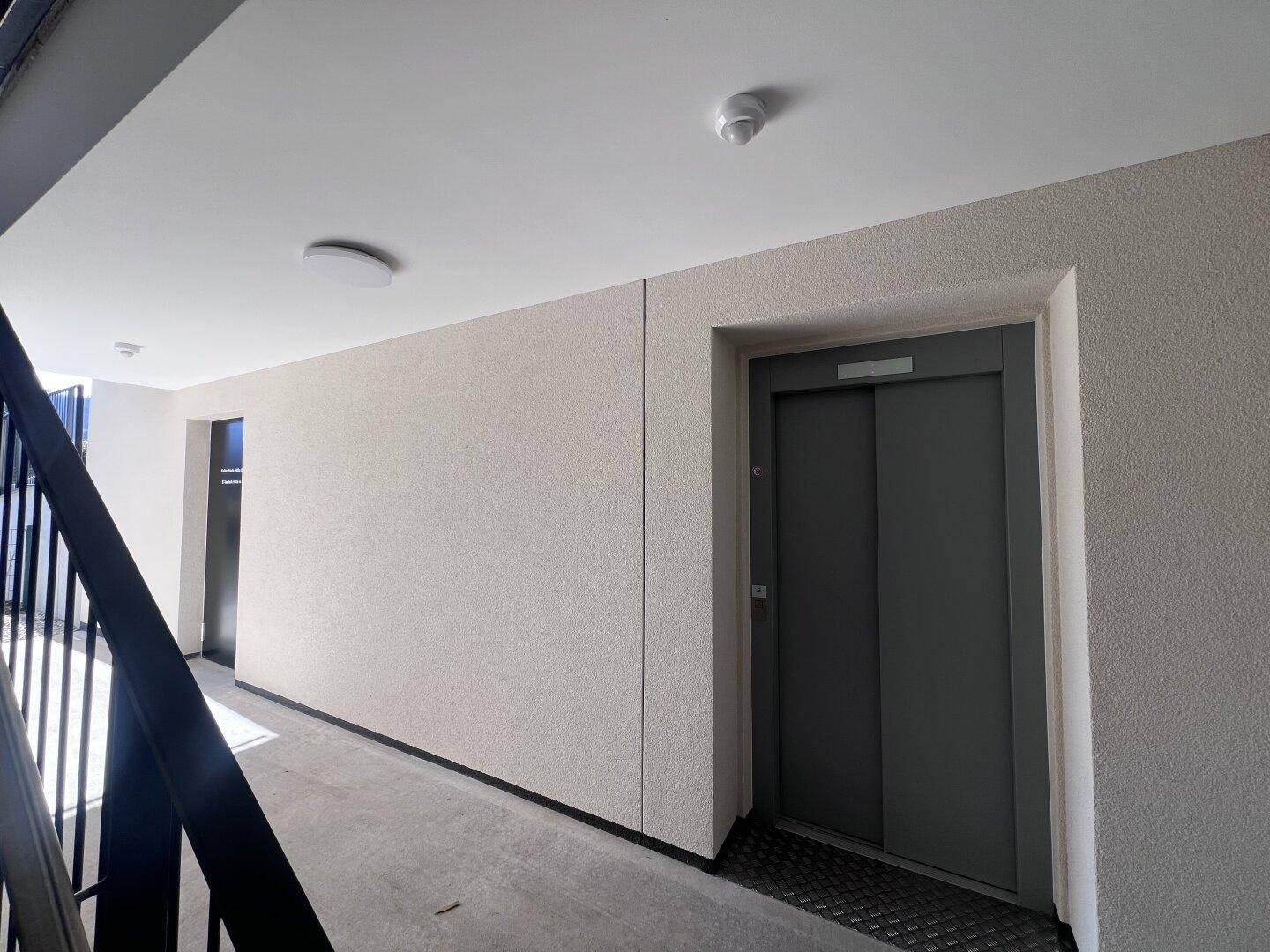 Heller Hausflur mit Aufzug und Treppenhaus, der zu den Wohneinheiten führt.