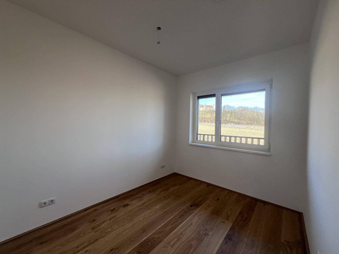 Unmöbliertes Zimmer mit Holzboden und Fenster, das einen weiten Blick ins Grüne bietet.