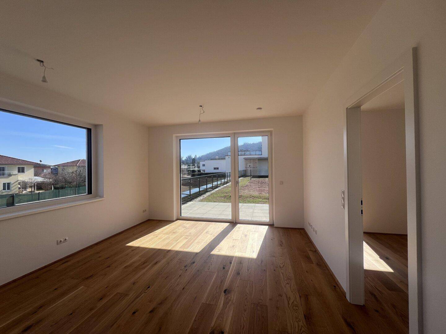 Geräumiger, heller Wohnraum mit hochwertigem Holzfußboden und großen Fenstern, die viel Tageslicht hereinlassen.