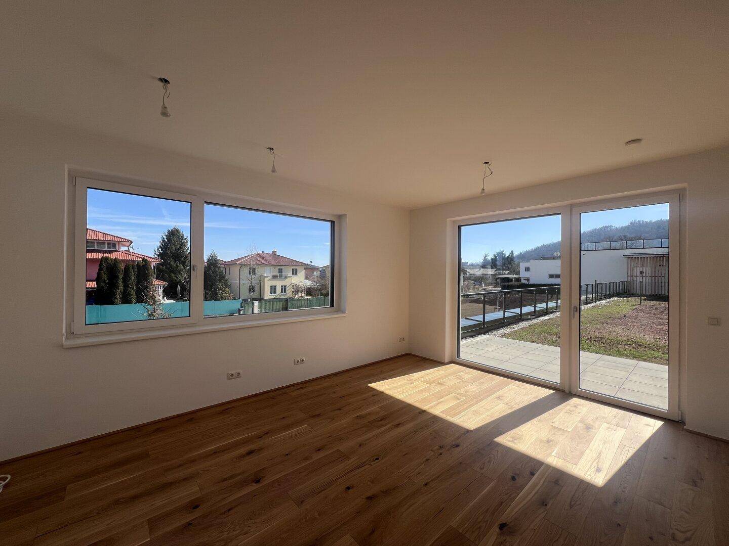 Heller, leerer Wohnraum mit neuem Holzfußboden, großen Fenstern und direktem Zugang zur Terrasse.