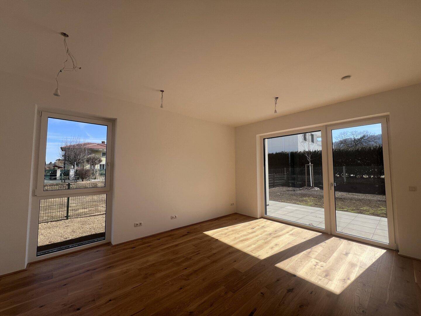 Heller Wohnraum mit Holzboden und großen Fenstern, die Zugang zur Terrasse bieten.