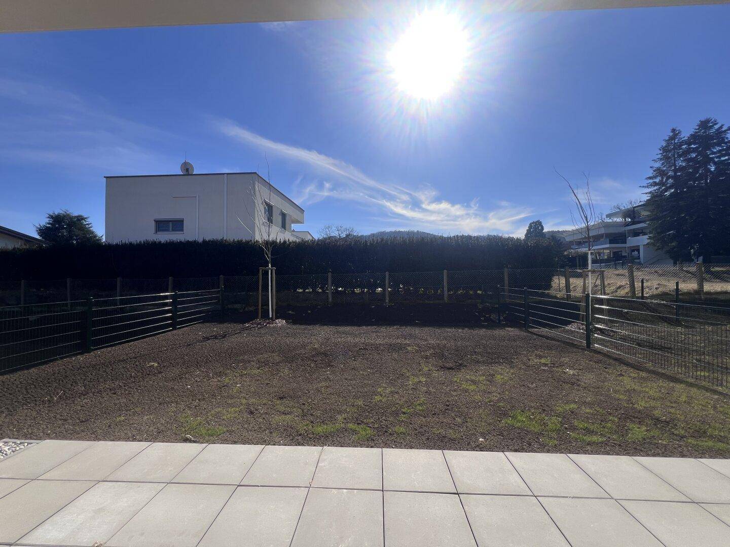 Sonnige Terrasse mit Fliesenboden und angrenzendem Gartenbereich vor einer modernen Wohnanlage.