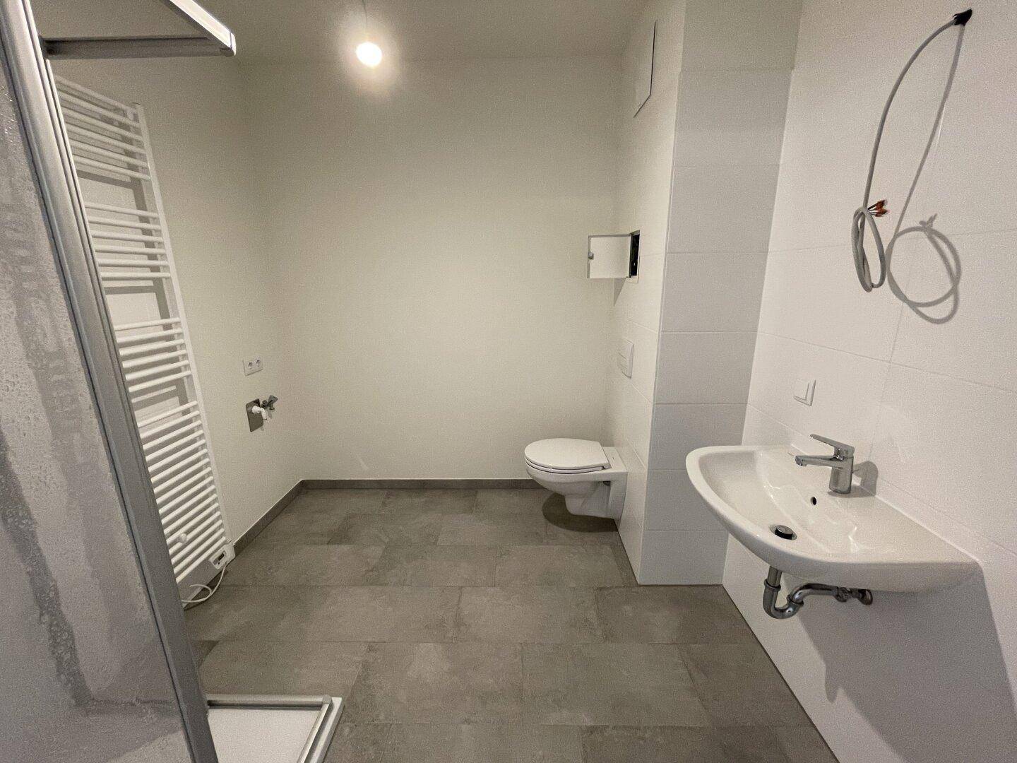 Modernes Badezimmer mit grauen Fliesen, wandhängendem WC, Einzelwaschbecken und Handtuchheizkörper.