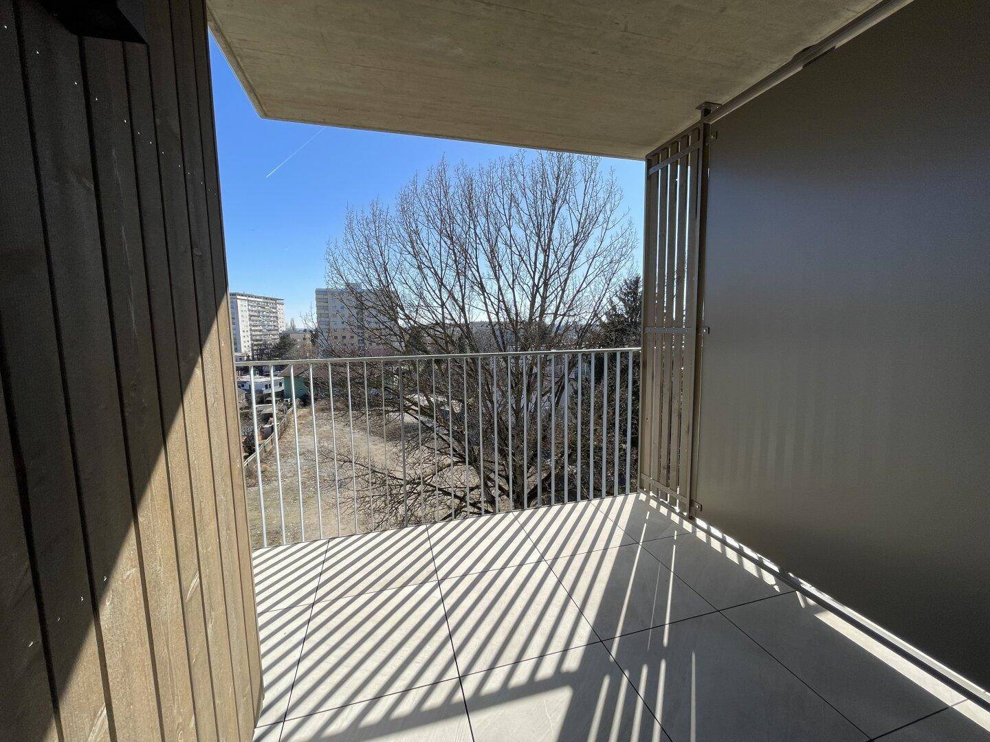 Sonniger Balkon mit Fliesenboden und seitlichem Blick auf Bäume und Gebäude.