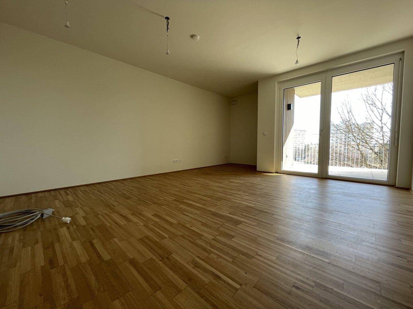 Geräumiges, leeres Wohnzimmer mit hellem Parkettboden und Zugang zum Balkon.
