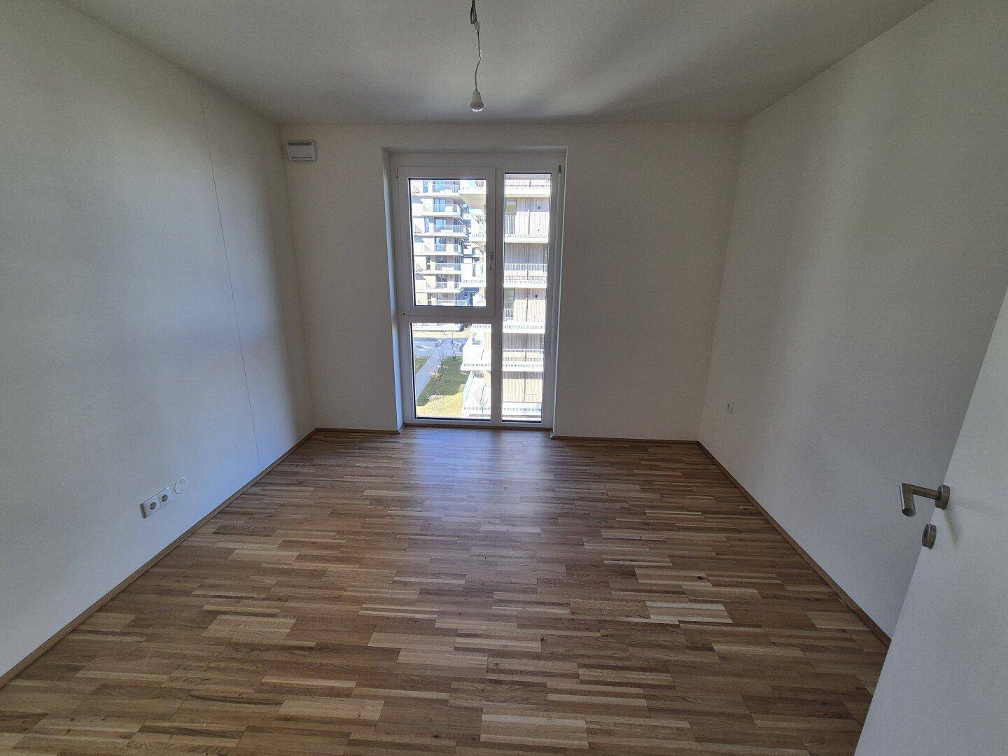 Helles Schlafzimmer mit Parkettboden und Fenster, das viel Tageslicht hereinlässt.