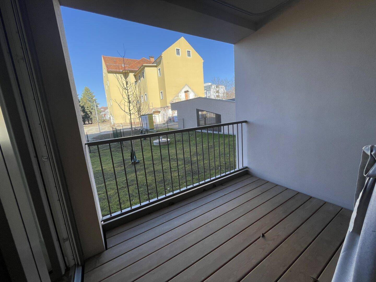 Sonniger Balkon mit Holzboden und Blick auf ein gelbes Gebäude und eine Rasenfläche.