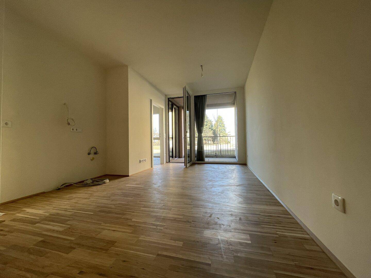 Heller, leerer Wohnraum mit Parkettboden und Zugang zu einem sonnigen Balkon.