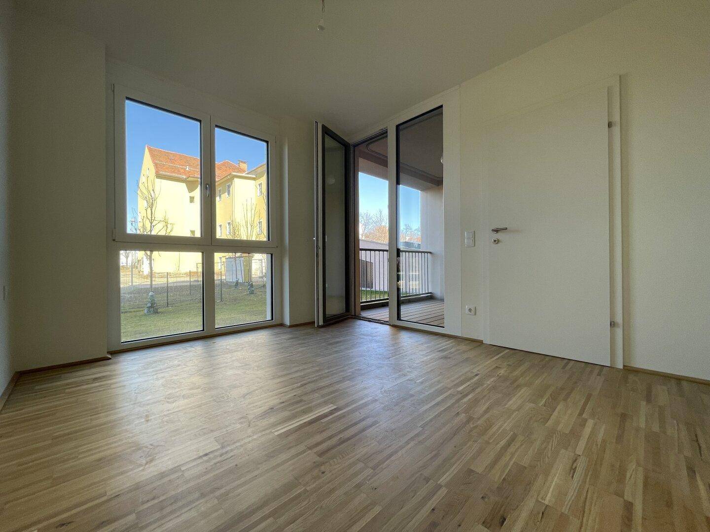 Lichtdurchfluteter Wohnraum mit großen Fenstern, Parkettboden und Zugang zum Balkon.