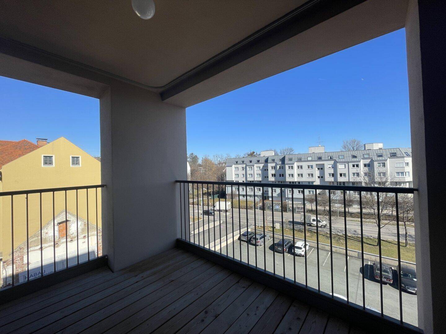 Heller Balkon mit Holzdielen und Blick auf die städtische Umgebung und Parkplätze.