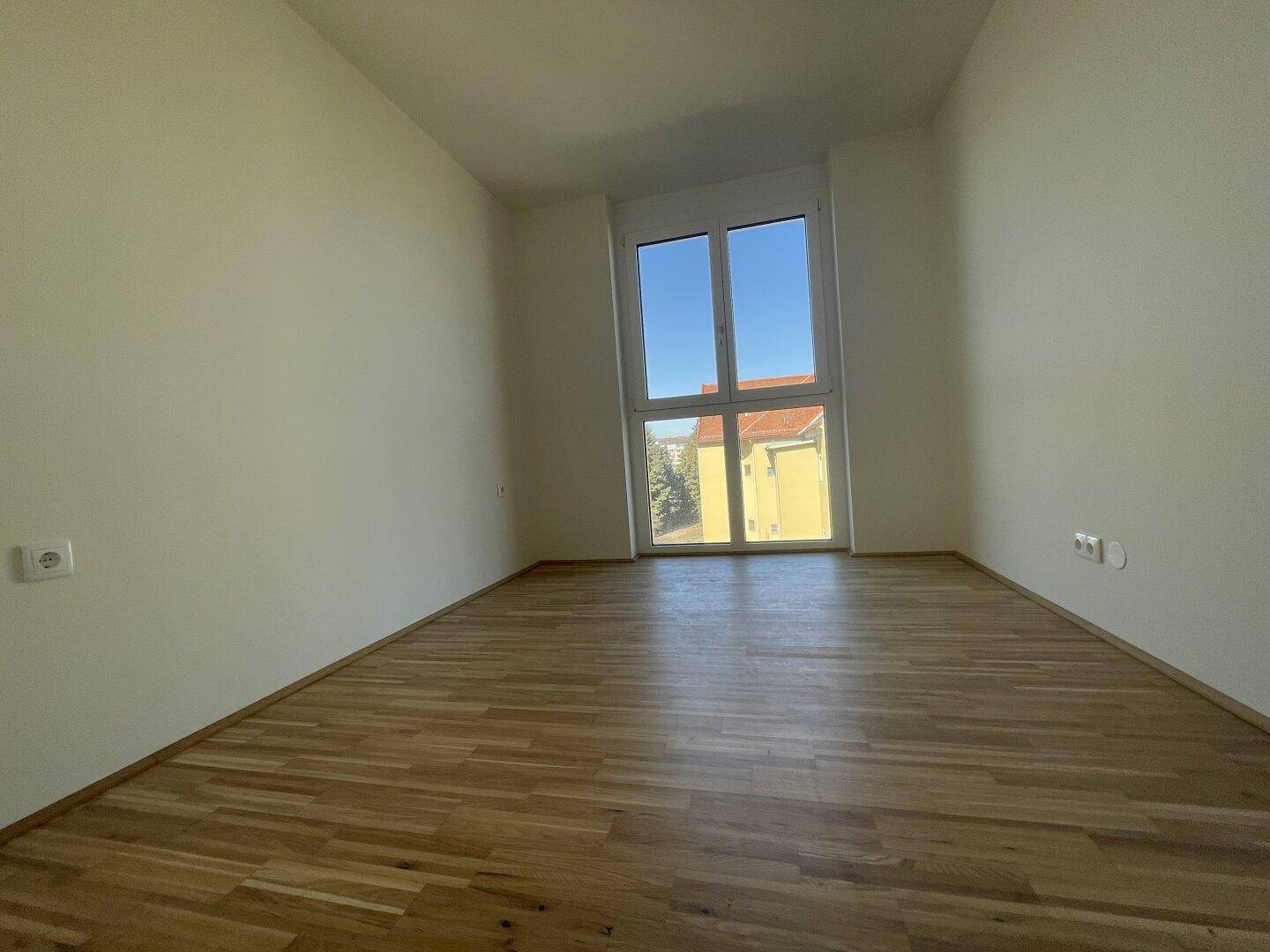 Helles Schlafzimmer mit Parkettboden und einem großen Fenster, das viel Licht hereinlässt.