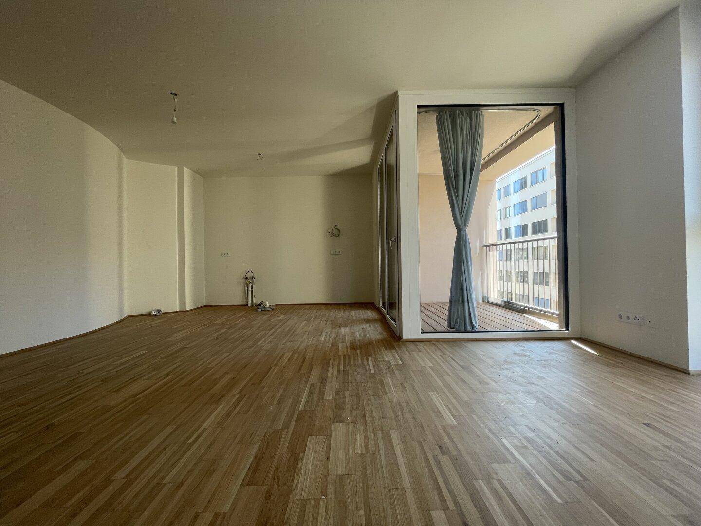 Geräumiges, leeres Wohnzimmer mit Parkettboden und Zugang zu einem sonnigen Balkon.