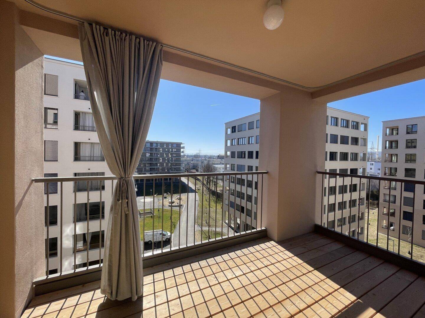 Heller Balkon mit Holzboden und Vorhang, bietet einen weiten Blick auf die Wohnanlage.