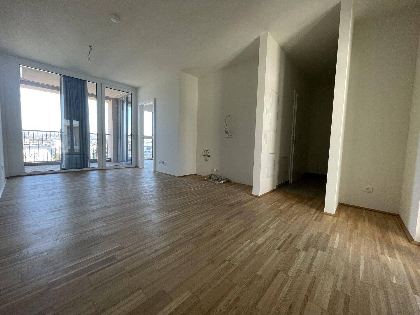 Geräumiges, leeres Zimmer mit Parkettboden und großen Fenstern zum Balkon.