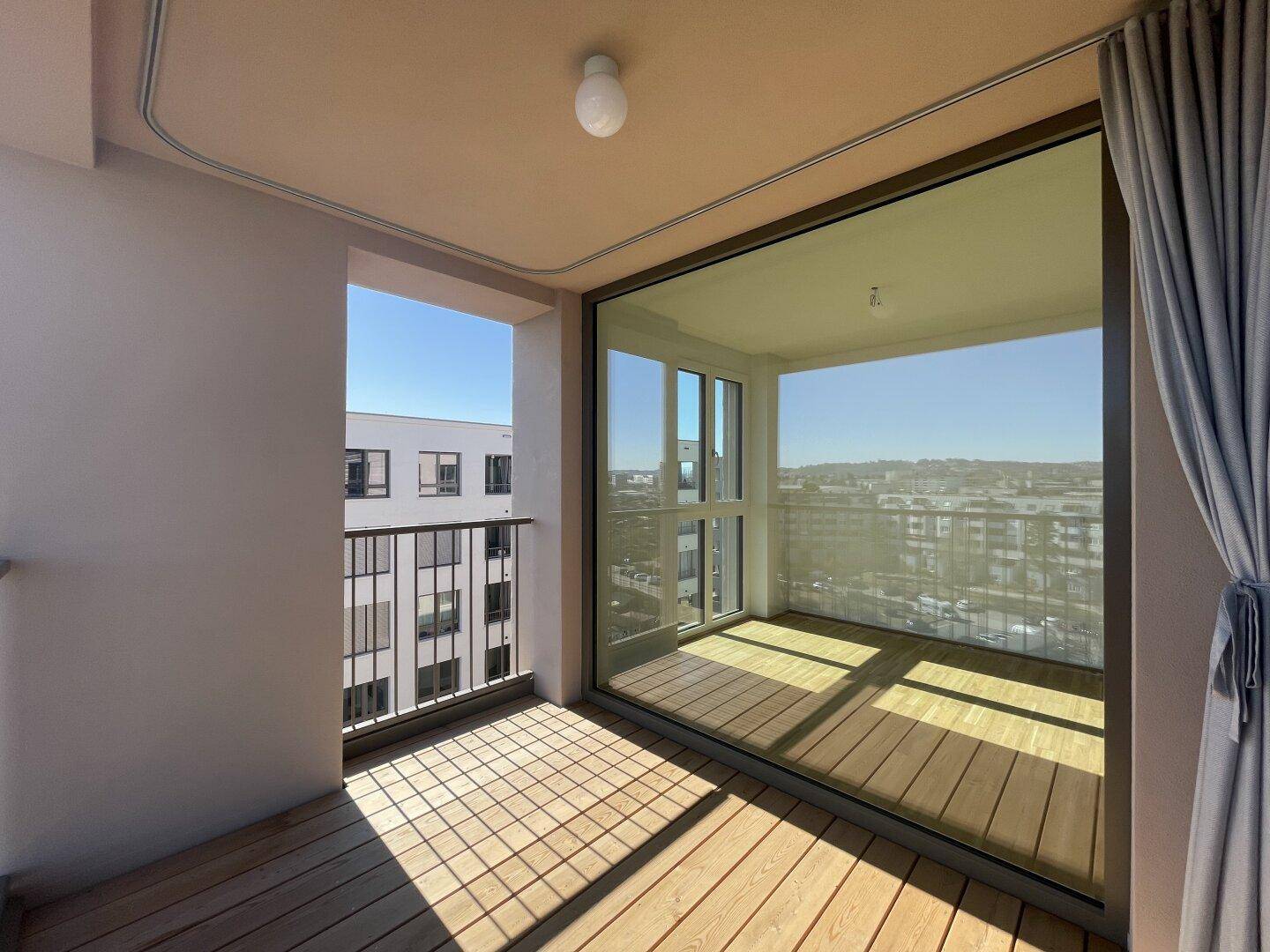 Sonniger Balkon mit Holzboden und Blick auf die umliegenden Gebäude und die Stadt.