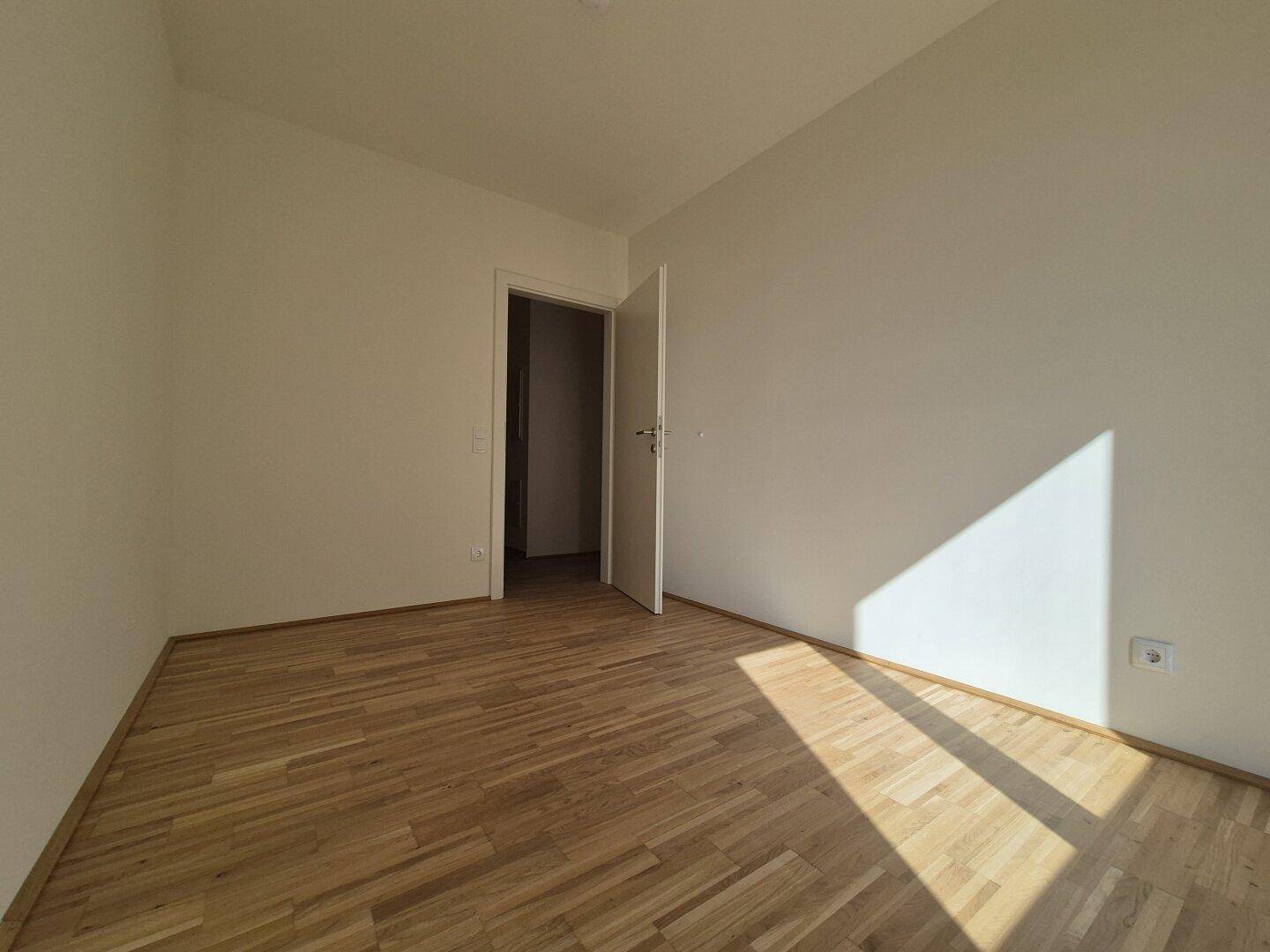 Helles, unmöbliertes Zimmer mit Parkettboden und weiß gestrichenen Wänden.