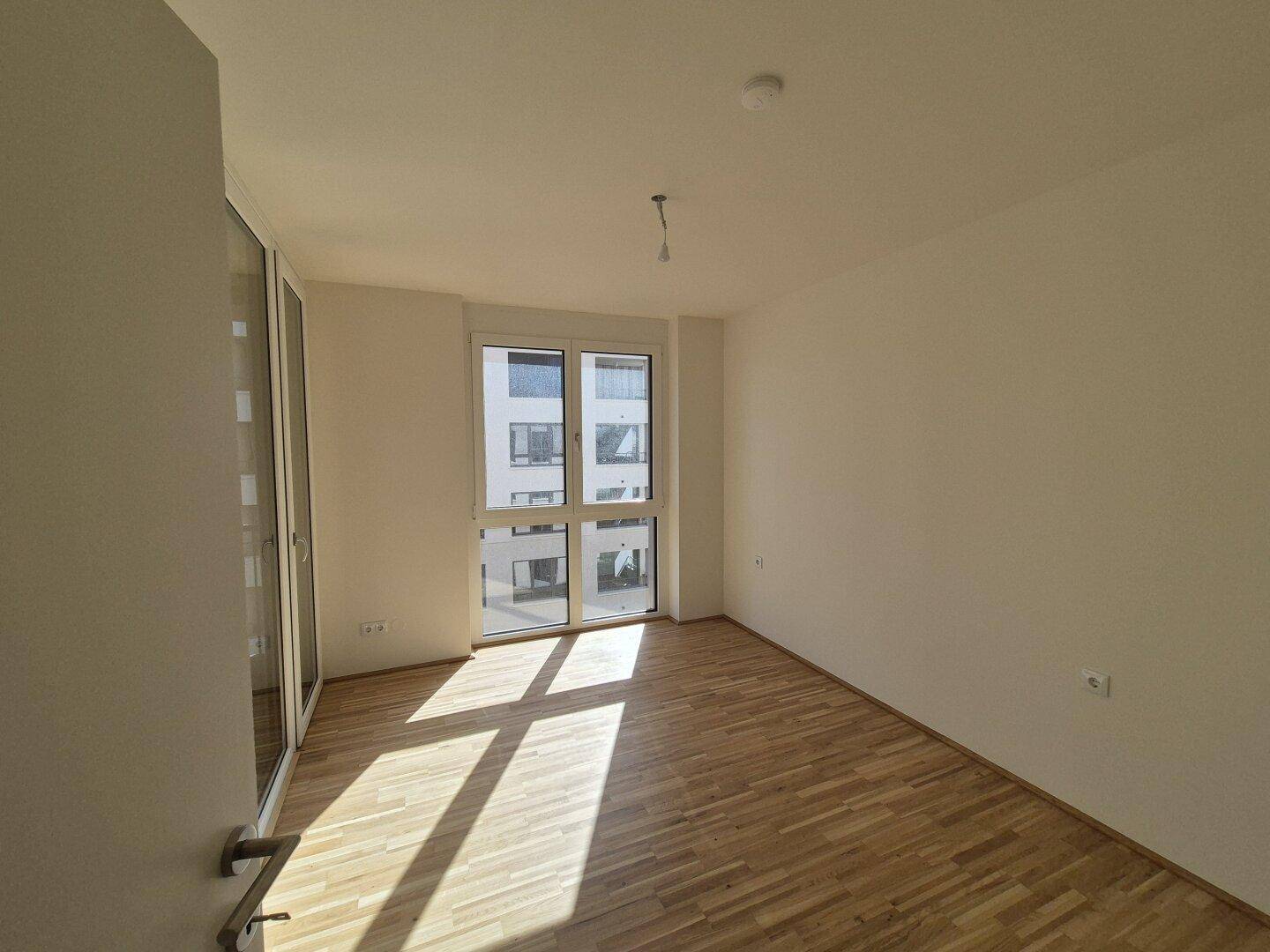 Lichtdurchflutetes leeres Zimmer mit Parkettboden und großem Fenster.