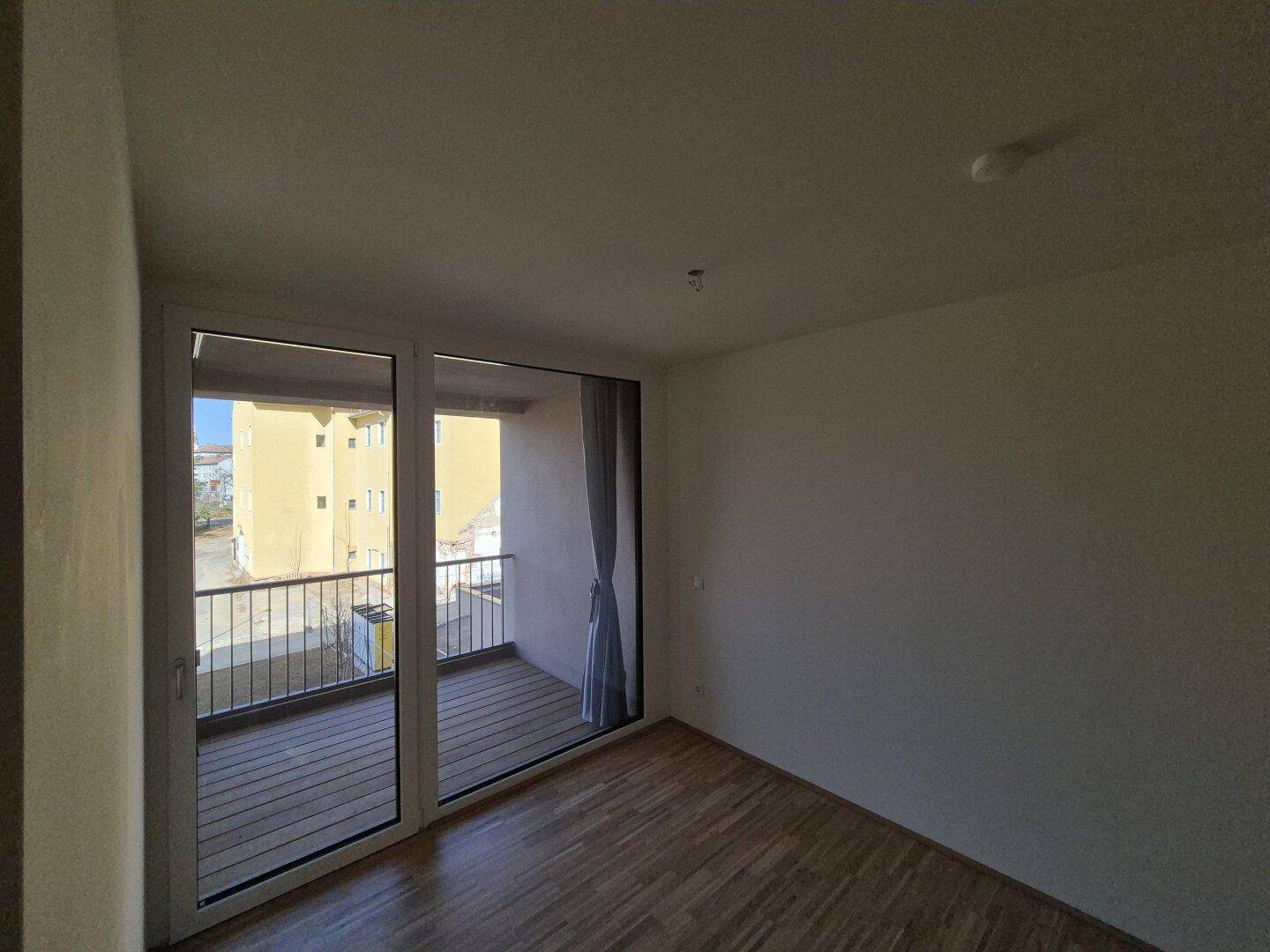Helles Zimmer mit Holzboden und großem Fenster mit Zugang zu einem Balkon.