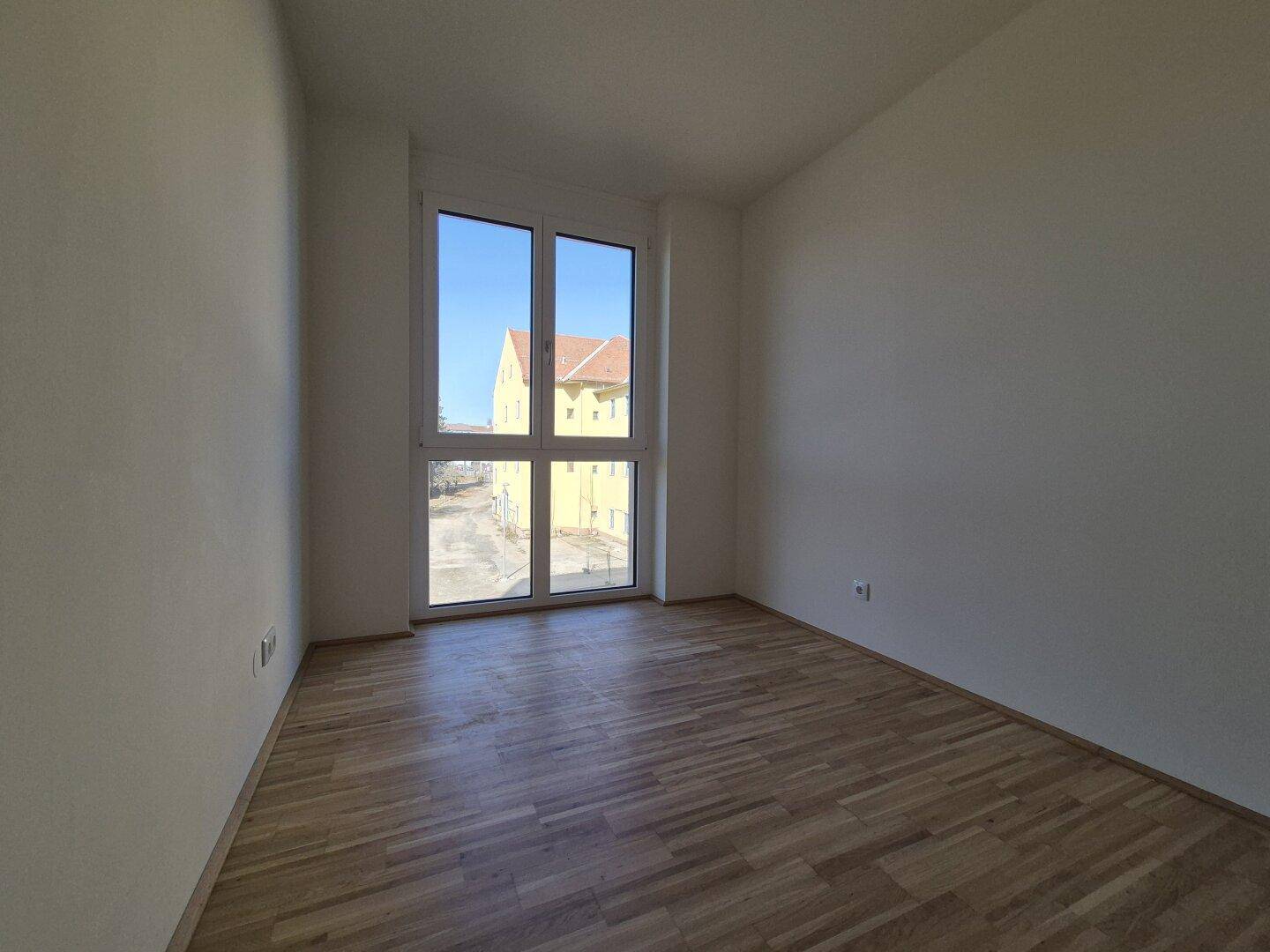 Helles Zimmer mit Holzboden und großem Fenster mit Blick auf die umliegenden Gebäude.