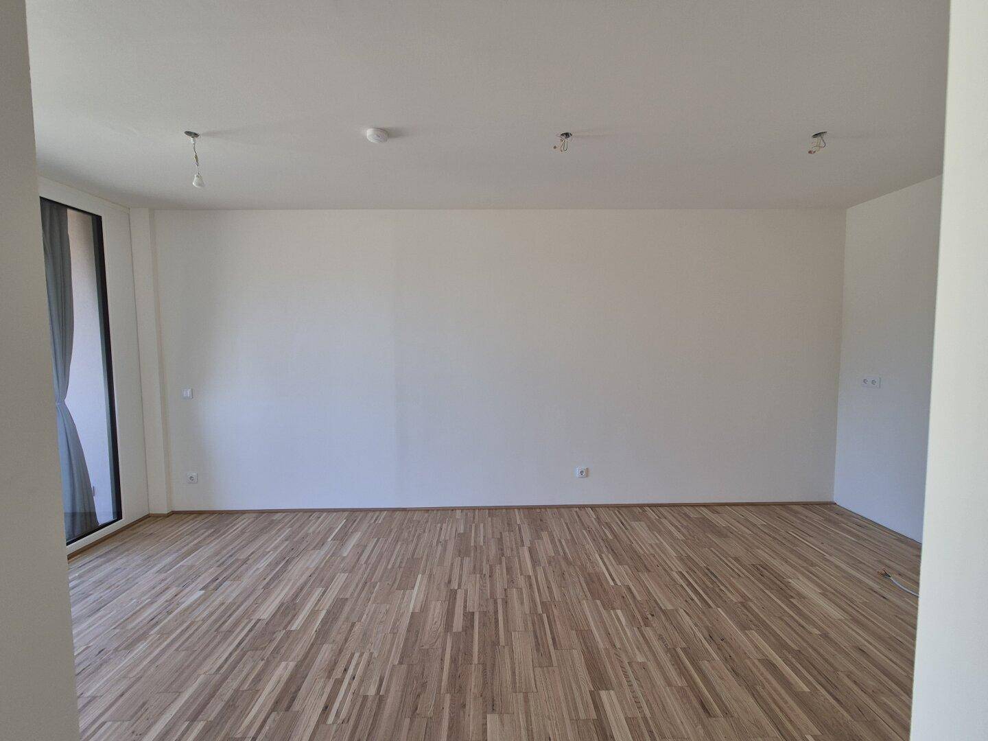 Geräumiges Wohnzimmer mit hellem Holzboden und großen Fenstern, die viel Licht hereinlassen.