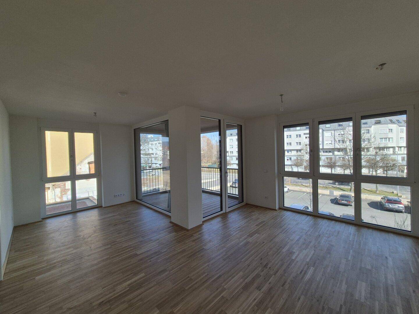 Heller, leerer Wohnbereich mit großen Fenstern und Zugang zum Balkon.