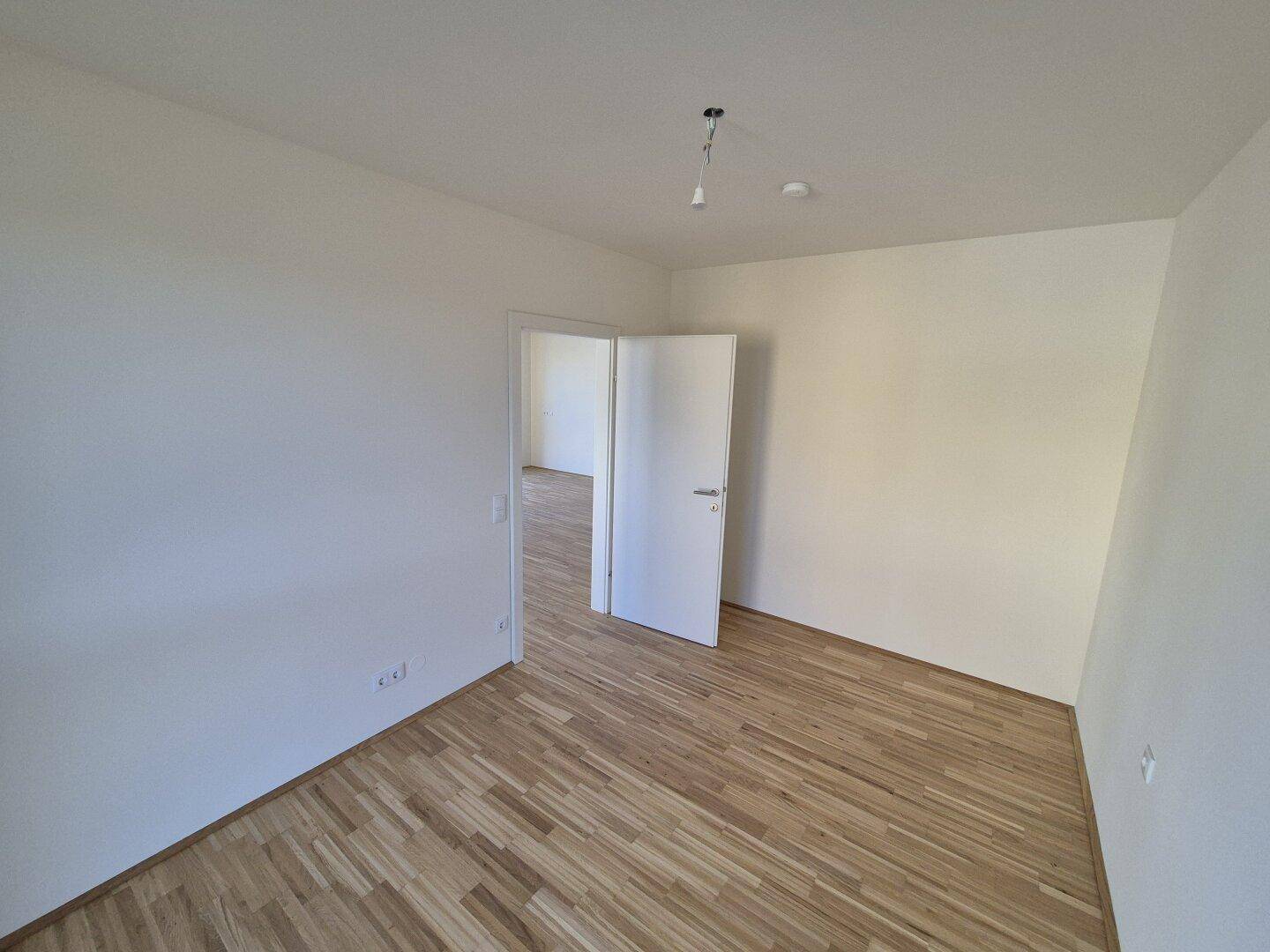 Leerer Raum mit Holzboden und weiß gestrichenen Wänden, mit offener Tür zu einem weiteren Raum.