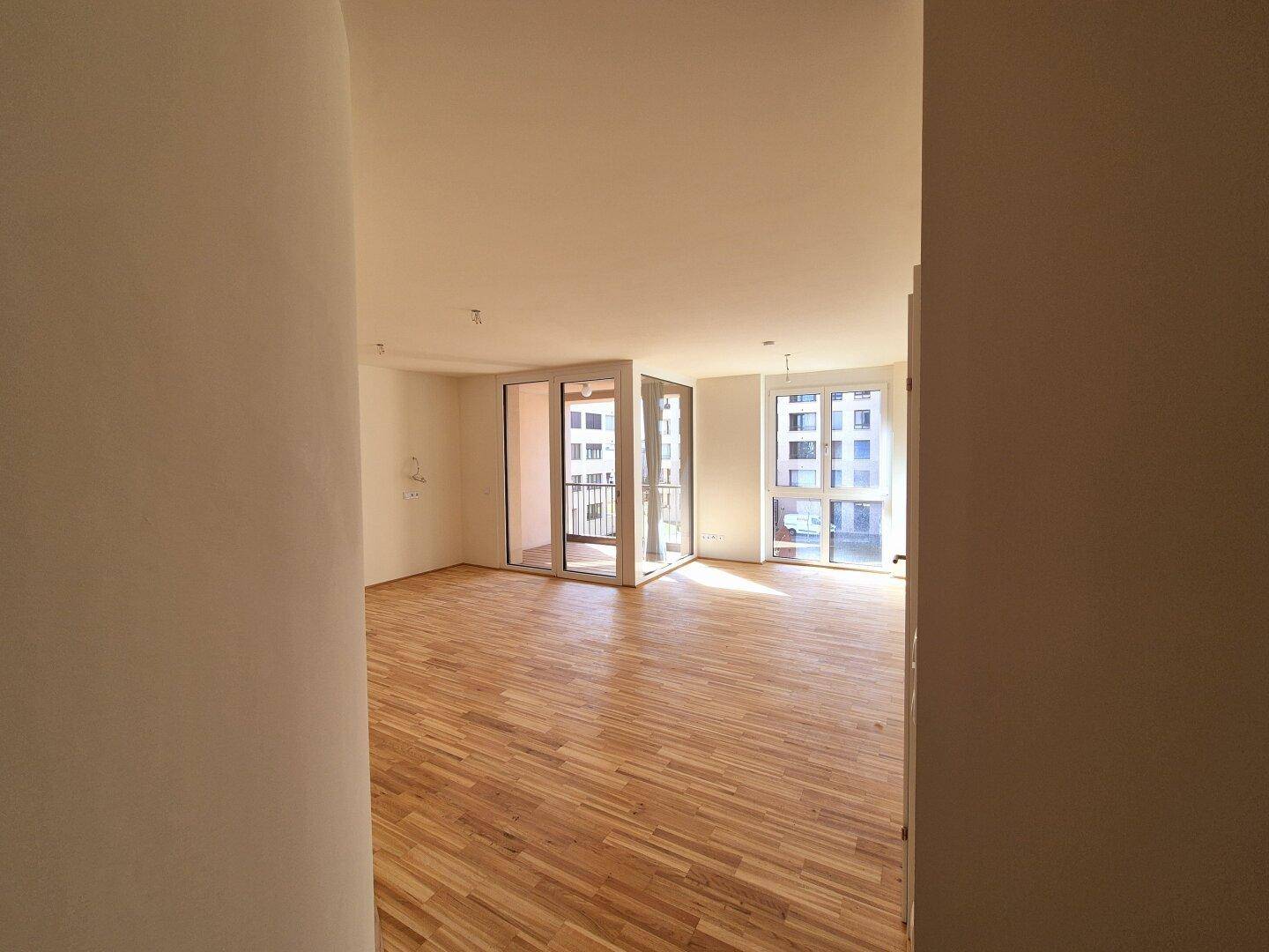 Geräumiges, leeres Wohnzimmer mit Parkettboden und Zugang zu einem sonnigen Balkon.