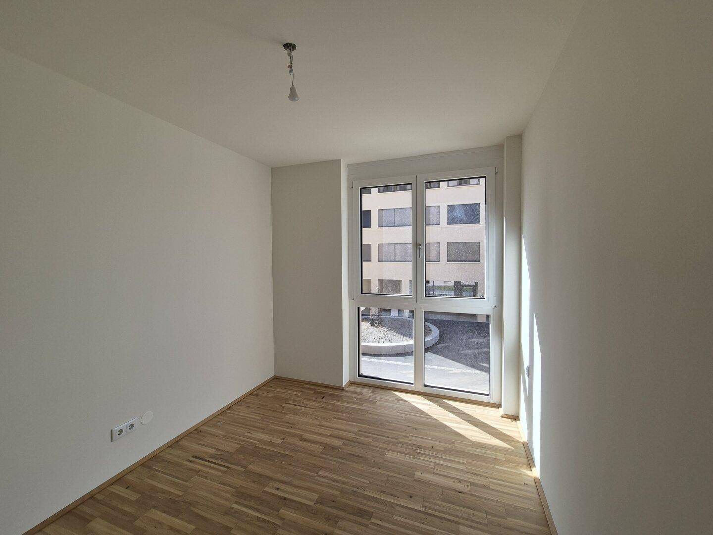 Helles Schlafzimmer mit Parkettboden und großem Fenster, das den Blick auf den Innenhof freigibt.