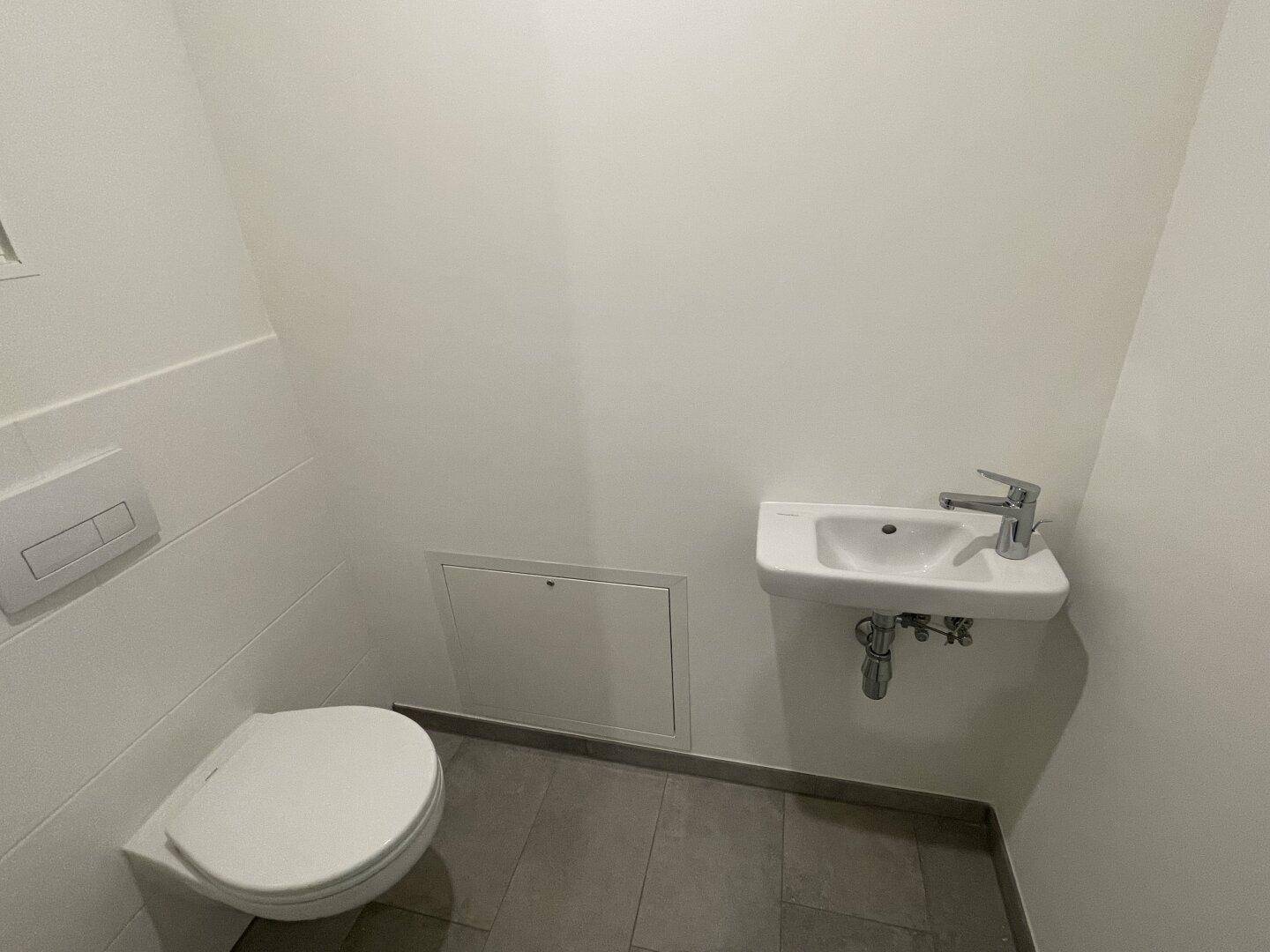 Modernes Gäste-WC mit Wand-WC, kleinem Waschbecken und grauen Fliesen in minimalistischem Design.