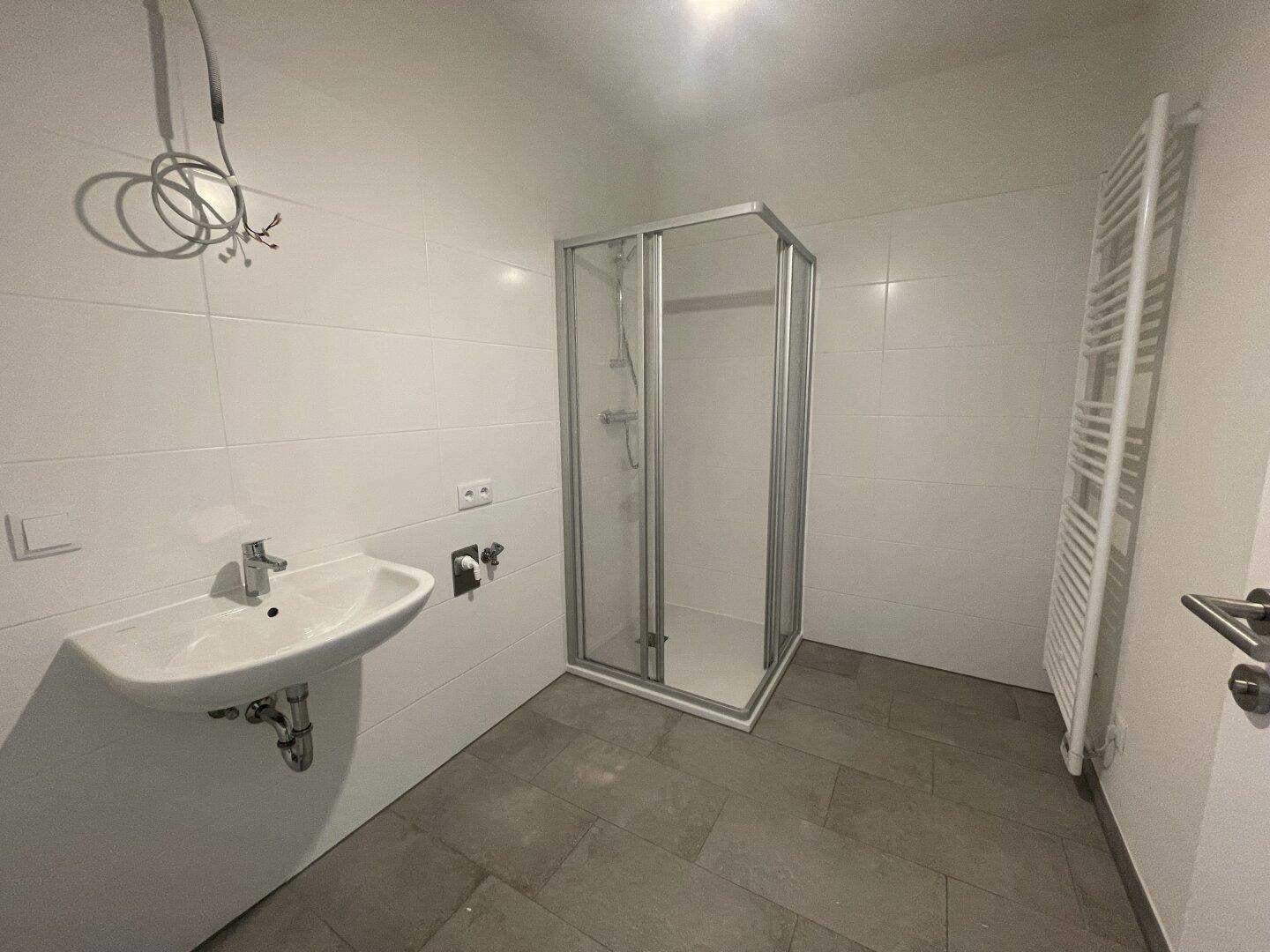 Badezimmer mit Duschkabine, Waschbecken, beheiztem Handtuchhalter und grauen Bodenfliesen.