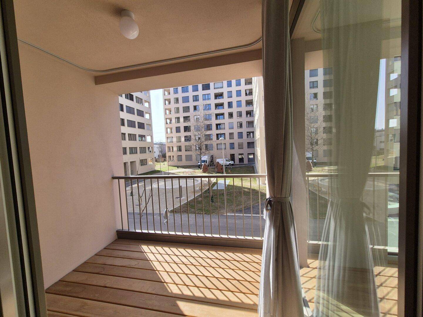 Sonniger Balkon mit Holzboden und Blick auf den Innenhof der Wohnanlage.