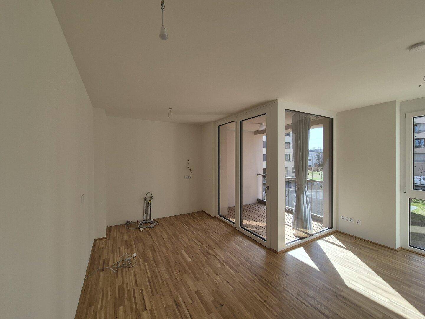 Geräumiges, leeres Wohnzimmer mit hellem Parkettboden und viel Tageslicht durch große Fenster.