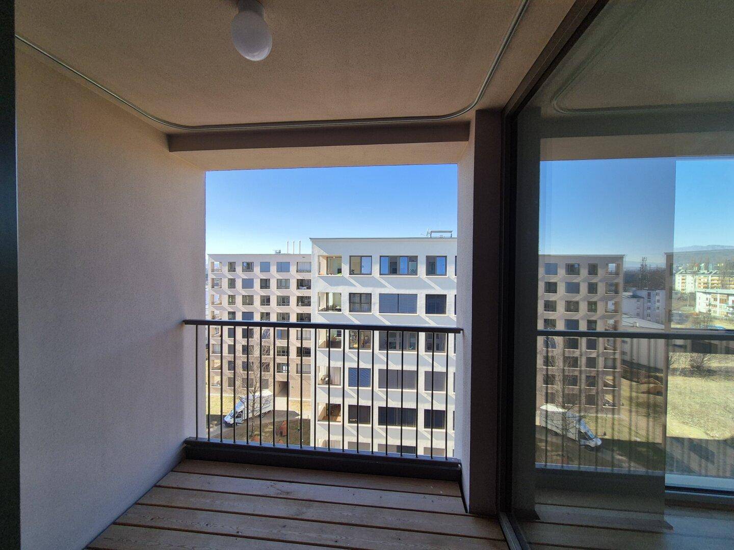 Heller Balkon mit Holzboden und Blick auf moderne Wohngebäude unter blauem Himmel.