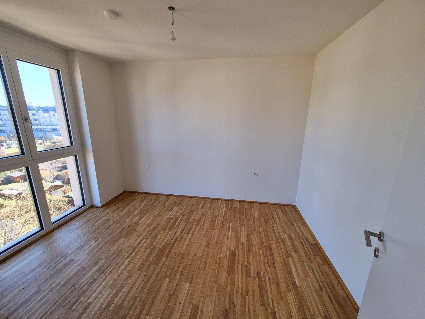 Helles Schlafzimmer mit Parkettboden und großem Fenster, das viel Tageslicht hereinlässt.